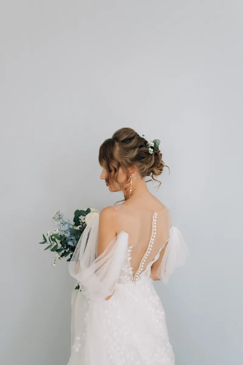 Elegant back view of a bride in a wedding dress holding a bouquet against a soft background.