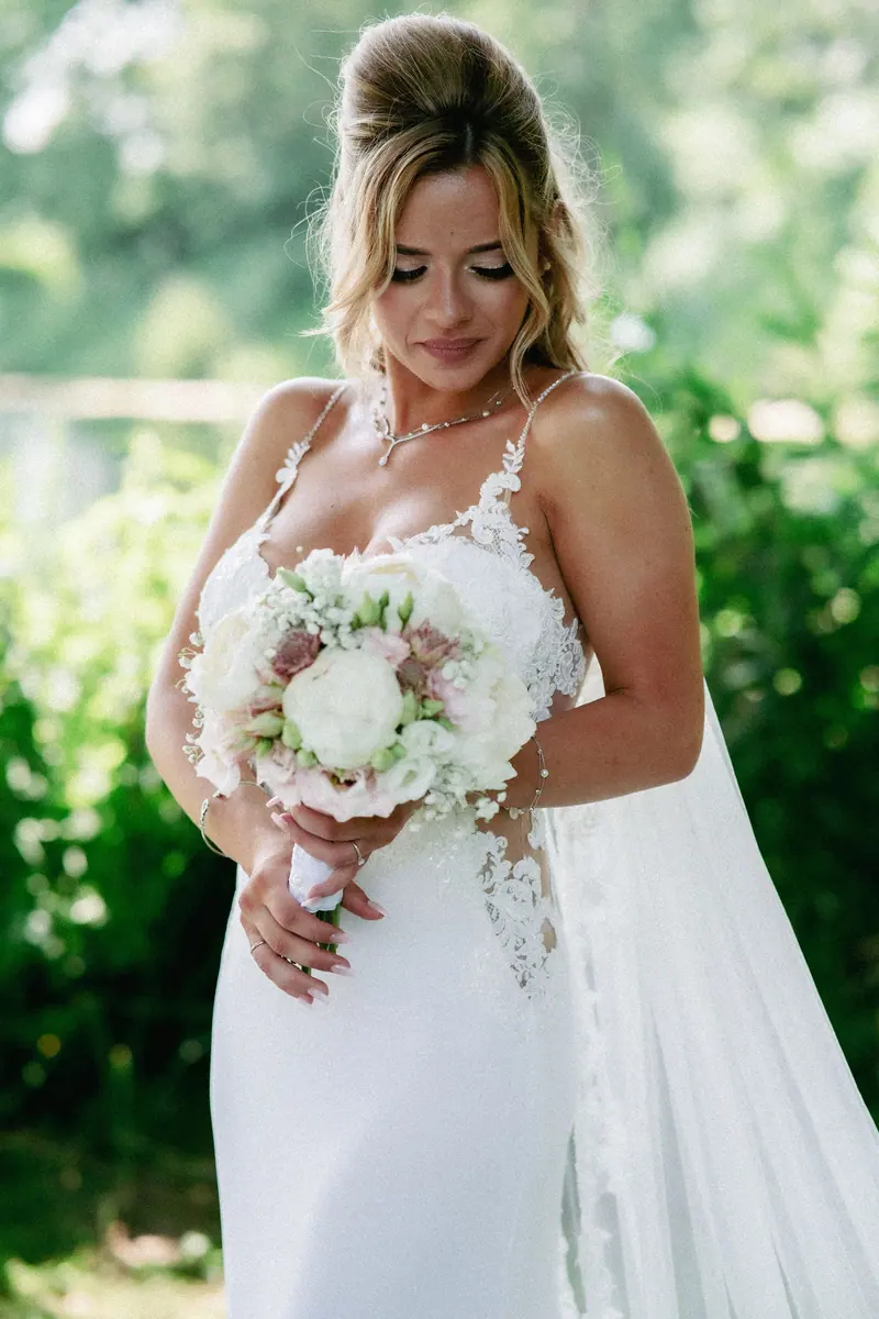 Beautiful bride in a lace wedding dress holding a bouquet amidst a lush green garden, capturing elegance and serenity.