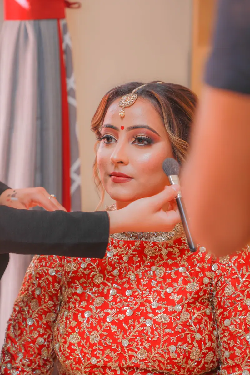 A woman in a red traditional dress having her makeup applied professionally indoors.