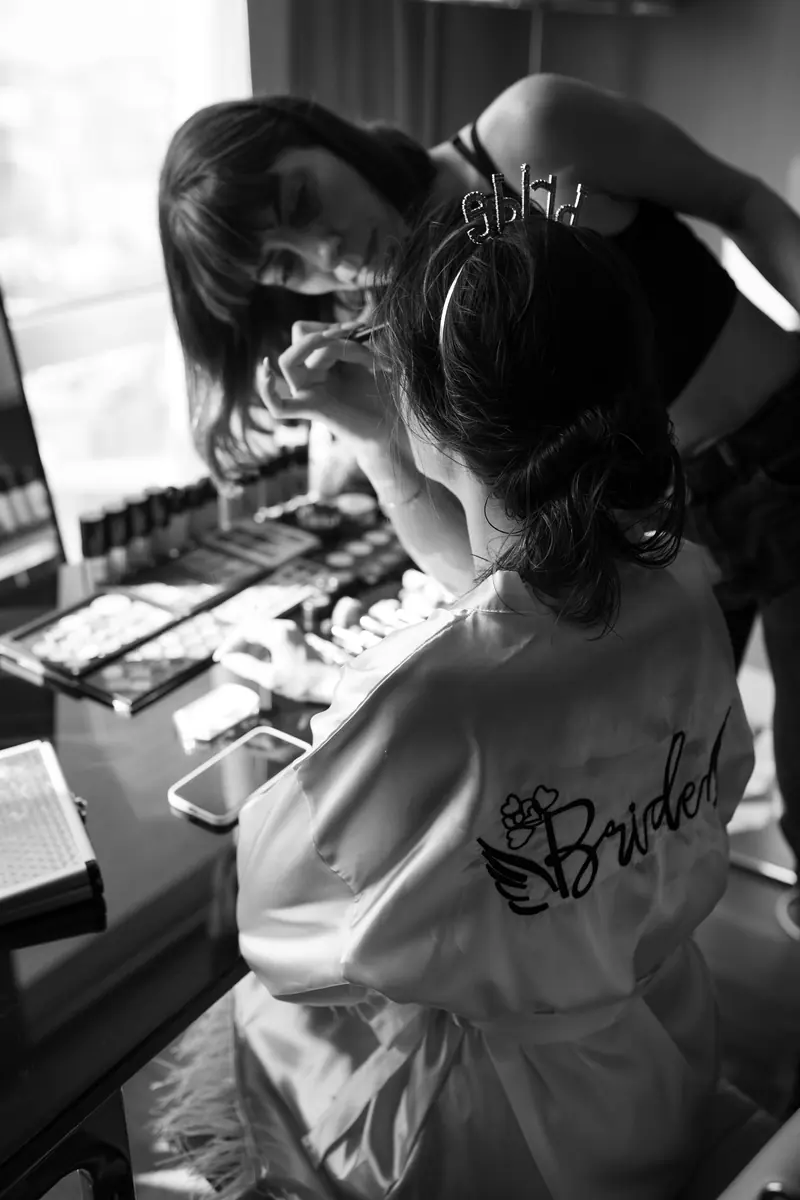 A bride gets her makeup done by a beautician, captured in a black and white setting.