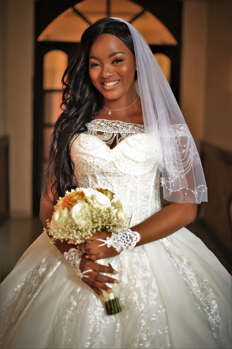 Elegant African bride in a stunning wedding dress holding a bouquet indoors, exuding joy and grace.