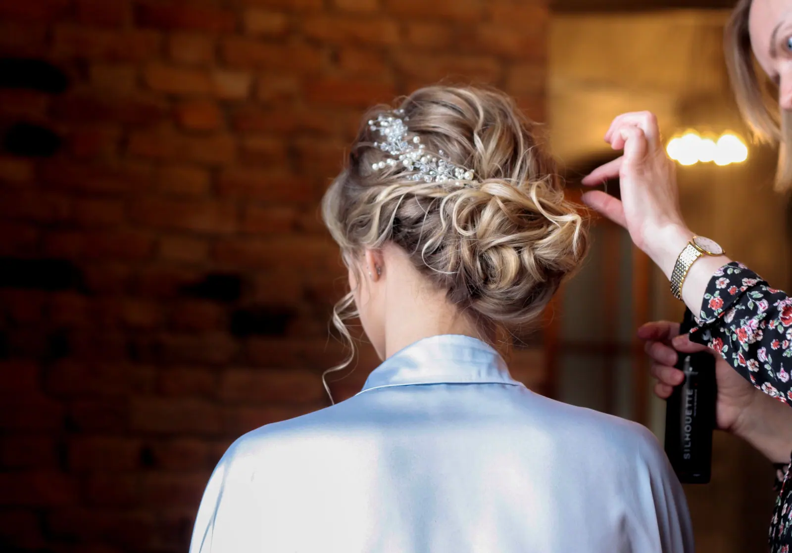 Stylist finishing a textured blonde bridal updo with a crystal comb in a warm brick-walled getting-ready room