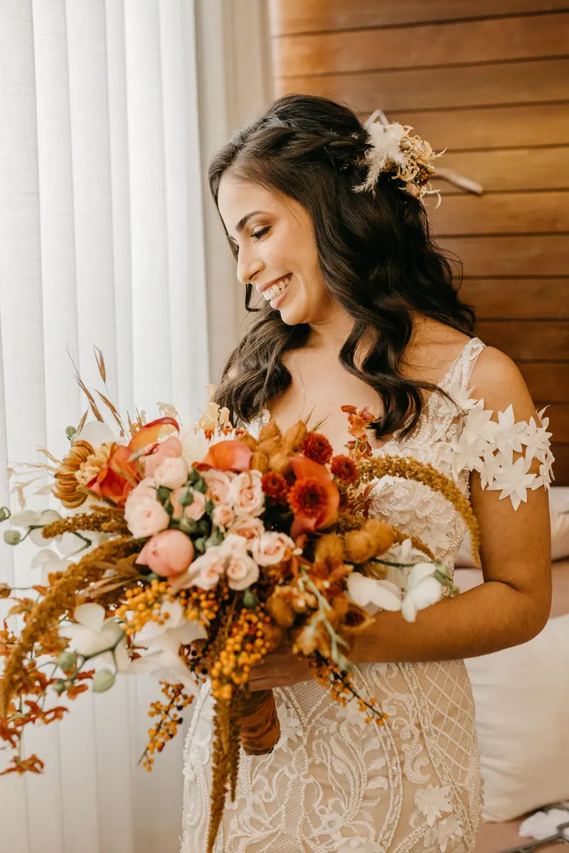 Bride in lace dress holding a stunning autumn-colored floral bouquet, smiling indoors.