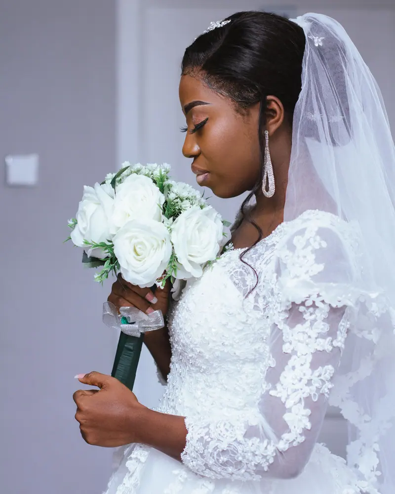 Beautiful bride in lace wedding dress holding a white floral bouquet indoors.