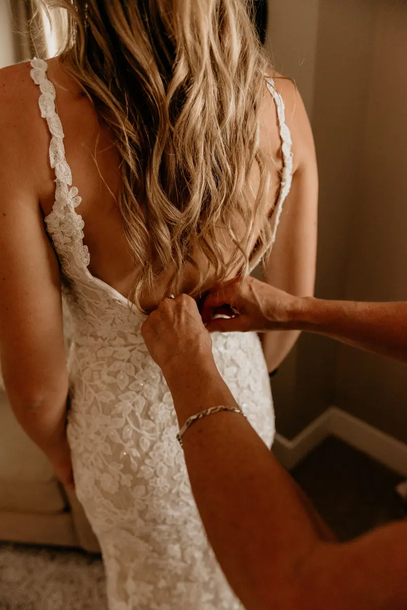 Back view of a bride in a lace wedding dress being zipped up, capturing the intimate getting-ready moment.