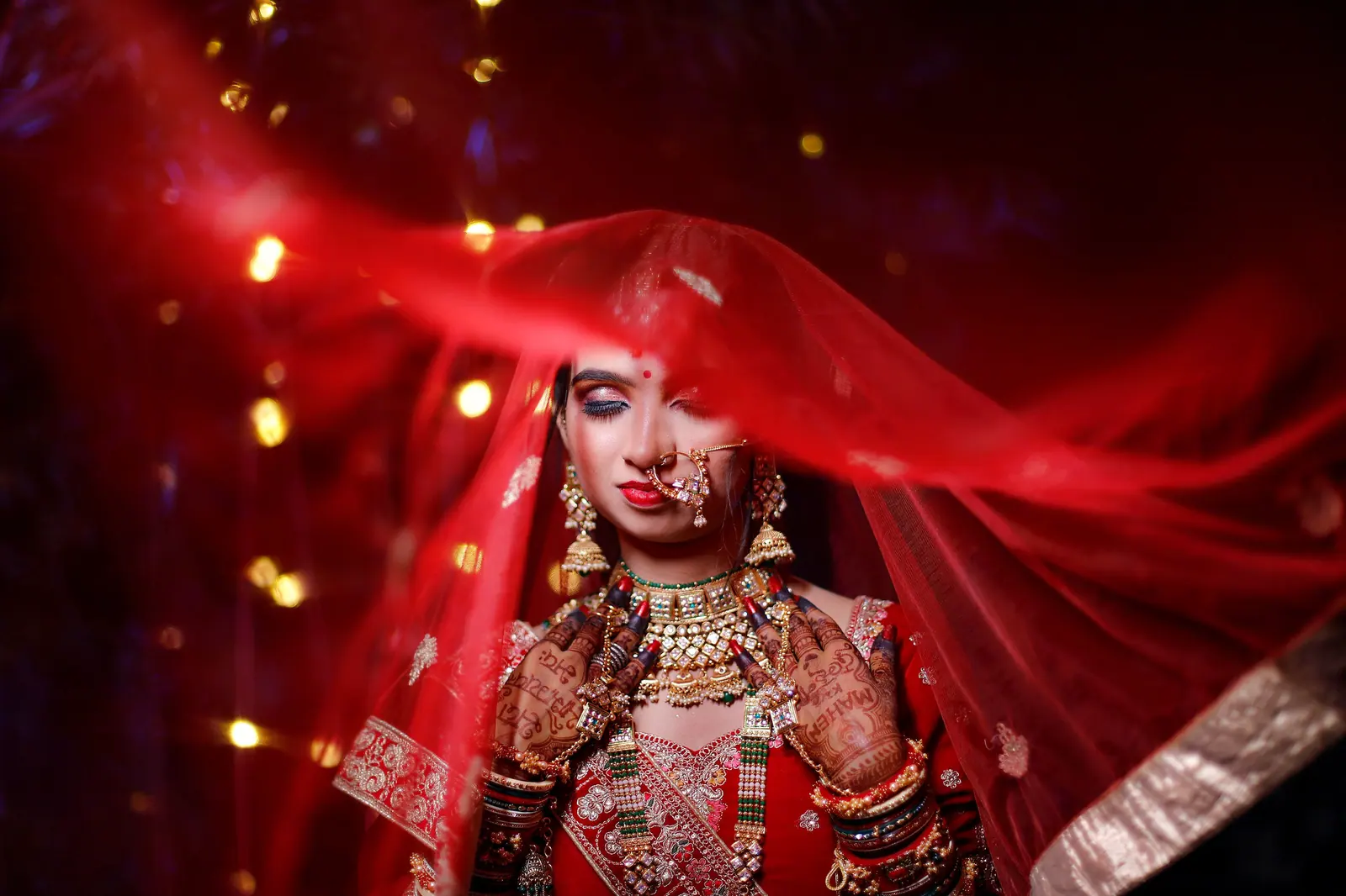 Bride in a red embroidered lehenga with a sheer red dupatta lifted around her face, glowing in warm light