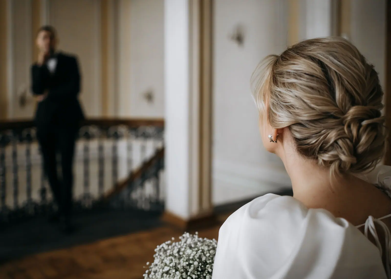 Bride with a soft textured low updo in a sunlit hotel suite while her groom waits on the staircase behind