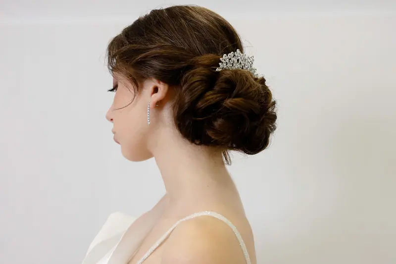 Side profile of a woman with an elegant bridal updo and sparkling tiara, set against a white background.