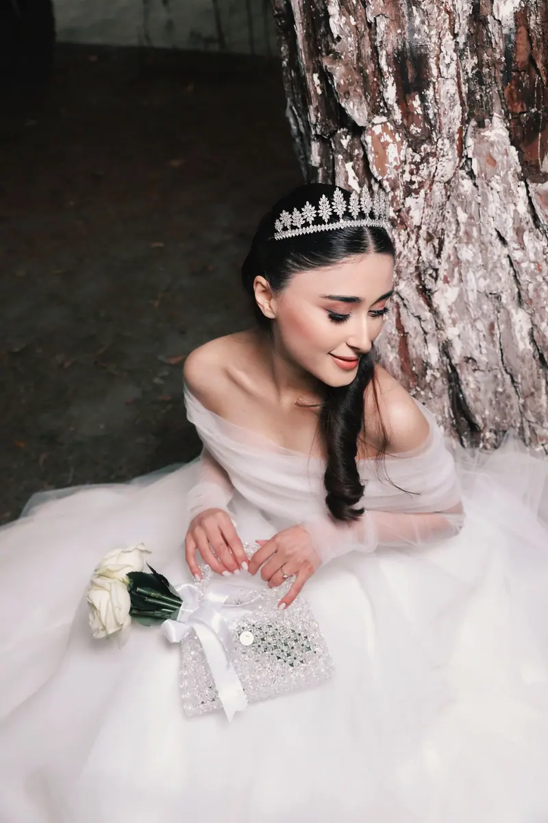 A beautiful bride in an elegant wedding dress sits gracefully by a tree, adorned with a floral tiara and holding a bouquet.