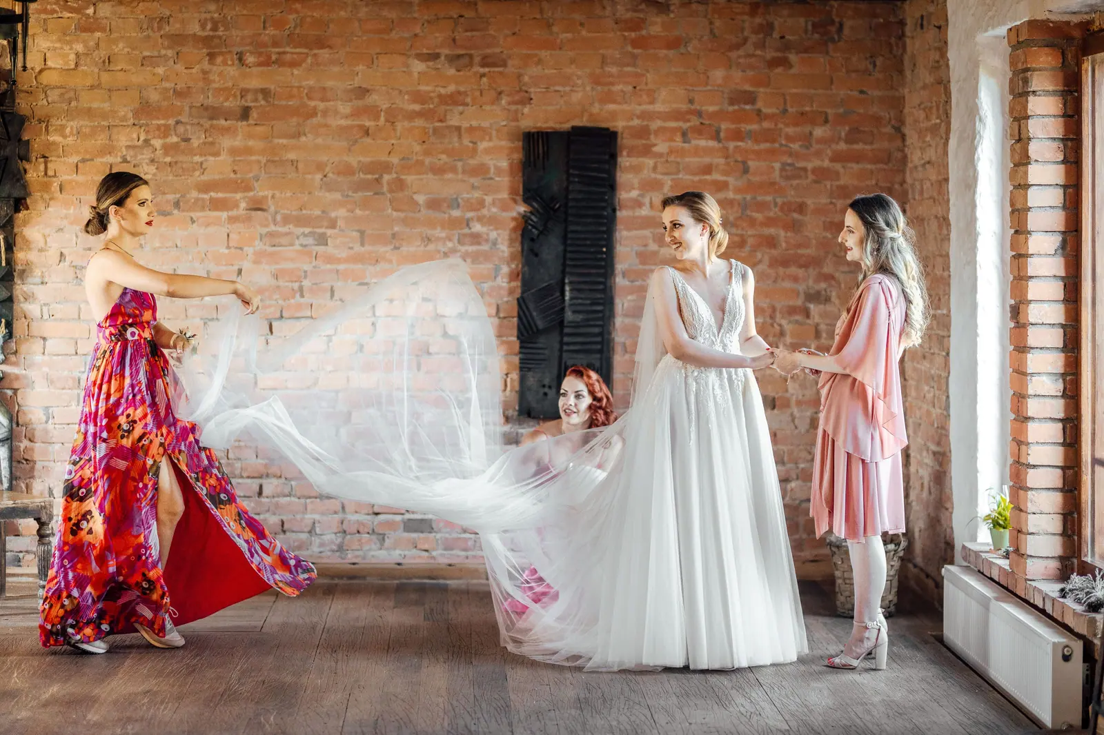 Bridal party enjoying a joyful moment indoors with vibrant, fashionable dresses and rustic backdrop.