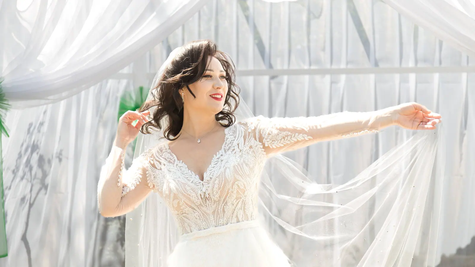 A bride in a lace wedding dress poses gracefully, holding her veil under a white tent.