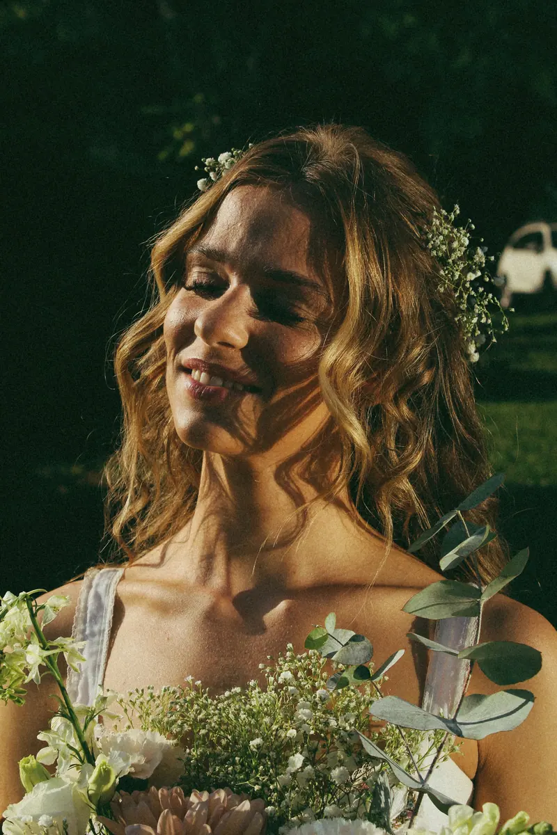 Radiant bride in a white dress holding a bouquet outdoors, basking in sunlight.