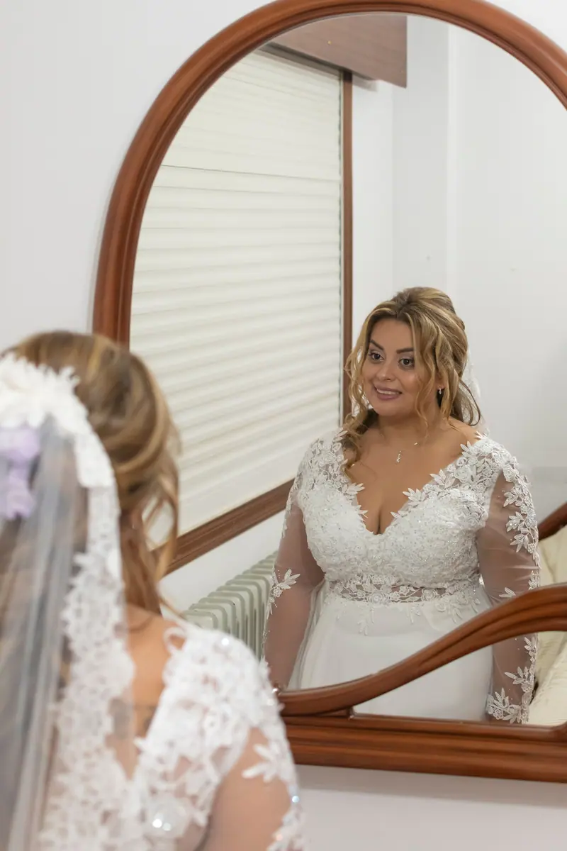 A smiling bride in a lace dress stands in front of a mirror, capturing a moment of joyful anticipation.