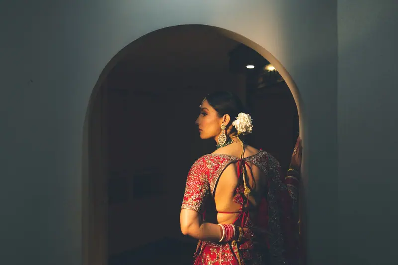 A beautiful Indian bride in a traditional red sari stands in a dimly lit doorway, showcasing intricate design and cultural beauty.