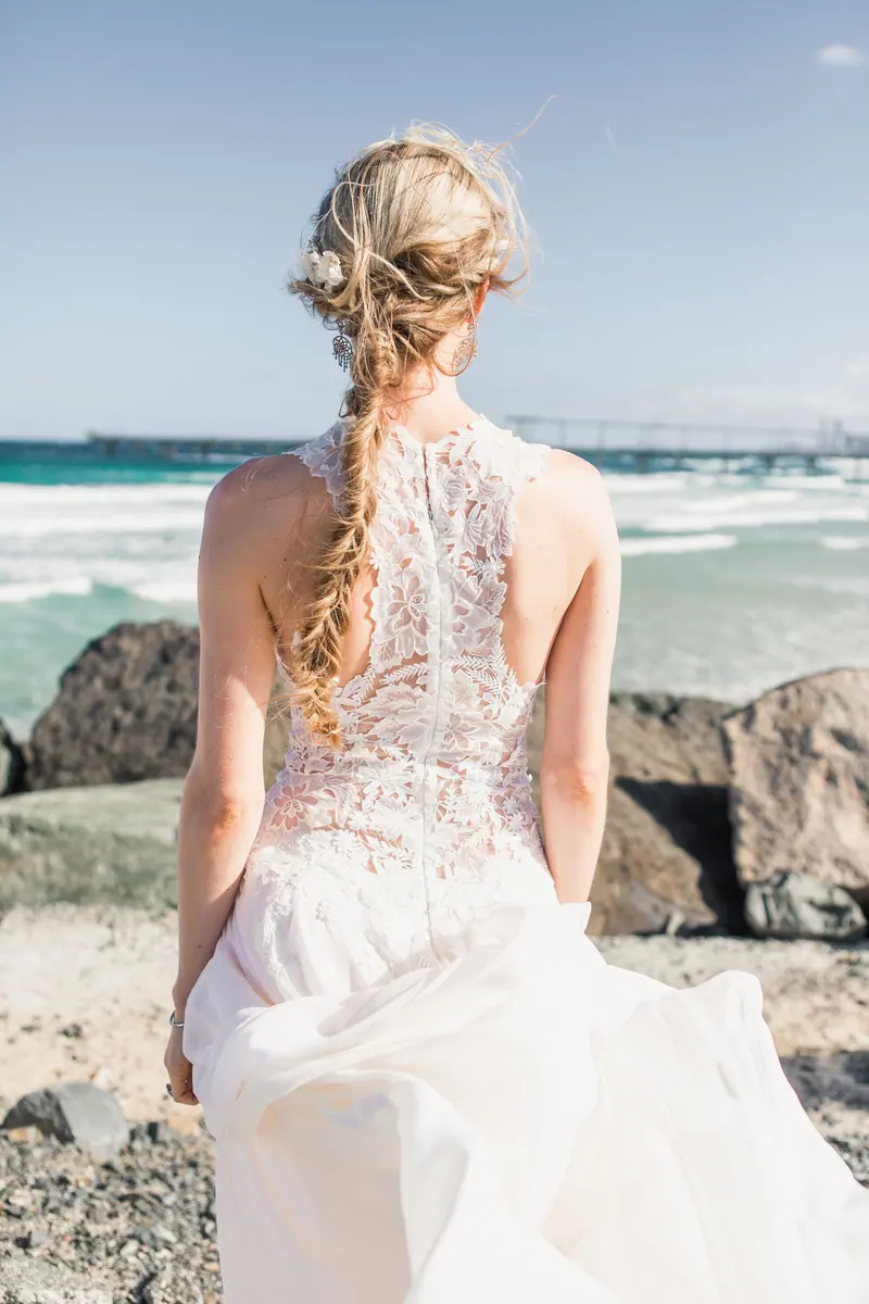 Back view of a bride in a lace wedding dress standing by the ocean with scenic waves.