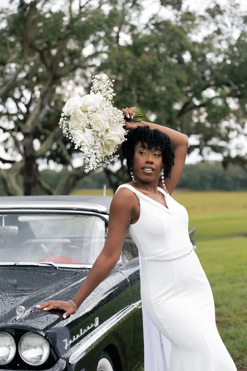 Beautiful bride in white dress posing with bouquet by vintage car outdoors.
