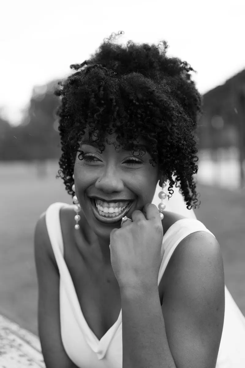 Black and white portrait of a joyful bride with curly hair and pearl earrings smiling outdoors.