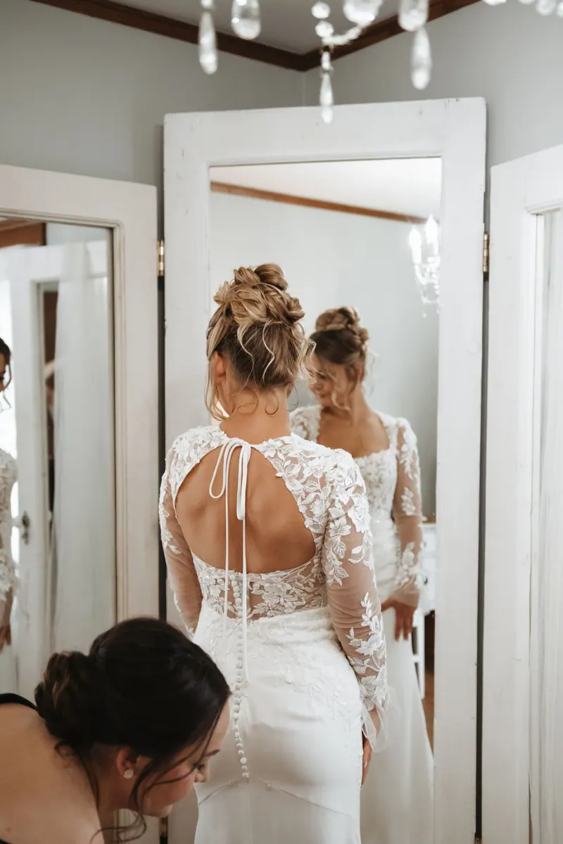 Bride in an elegant lace wedding gown getting ready with assistance indoors, surrounded by mirrors.