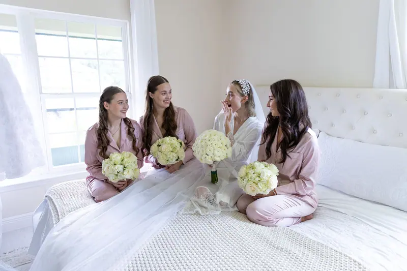 Bride and bridesmaids in pastel pajamas with bouquets, sharing a joyful moment before the wedding.