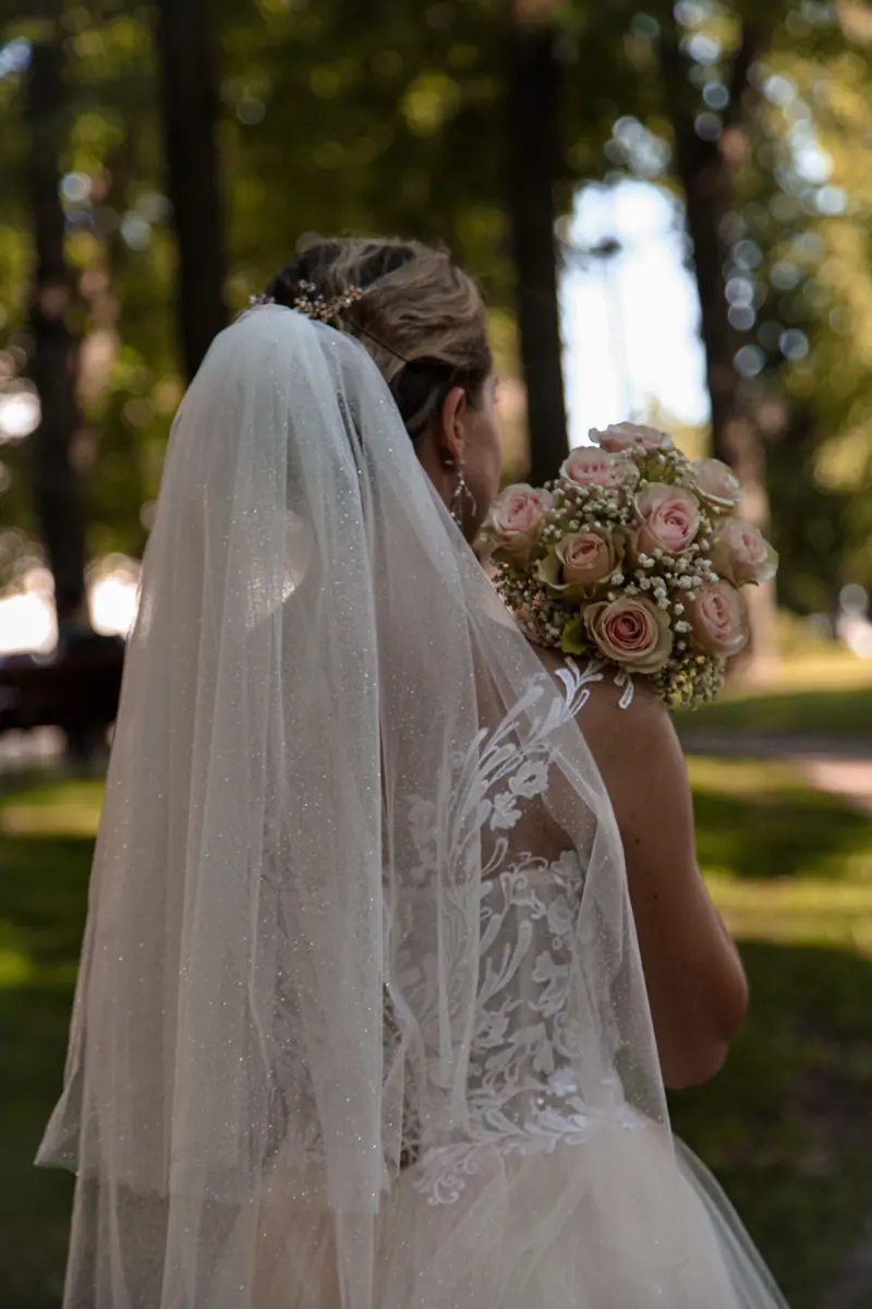 Bride in elegant dress holds bouquet in sunlit forest, captured from behind.