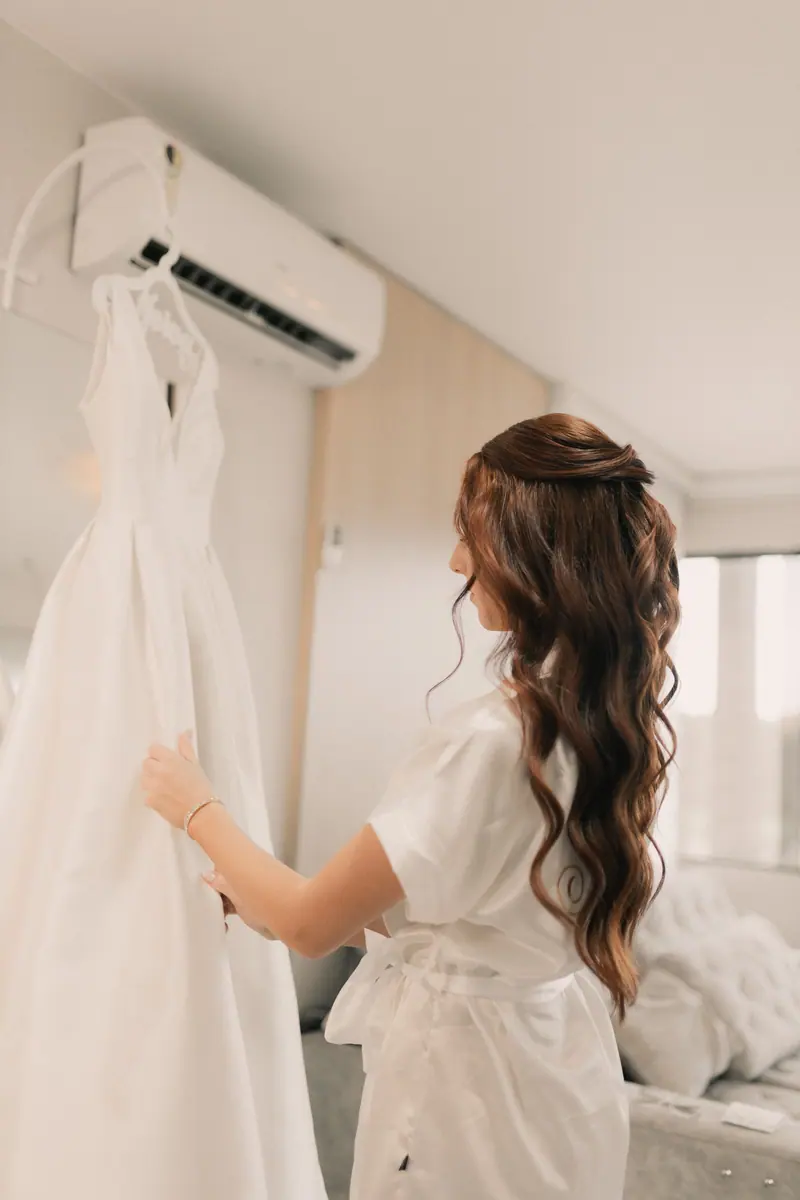 Bride gazing at her elegant white wedding dress hanging in a bright, serene room.