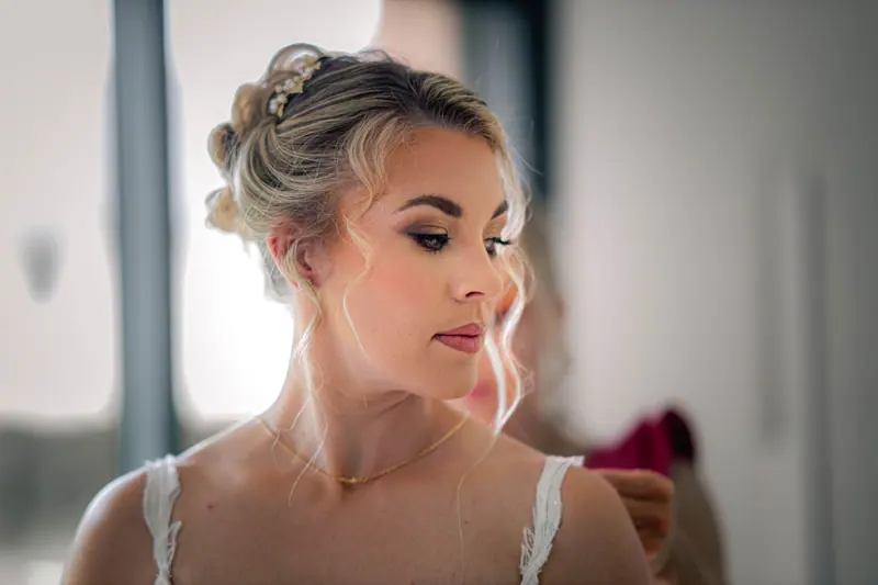A bride with styled blonde hair and makeup gets ready for her wedding in a bright room.
