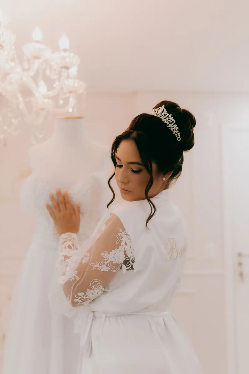 A bride in a lace robe and tiara admires a wedding dress, creating a serene bridal preparation scene.