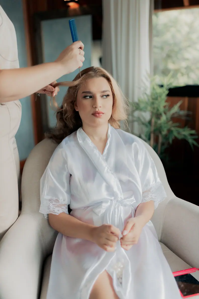 A woman in a white robe having her hair styled by a professional indoors.