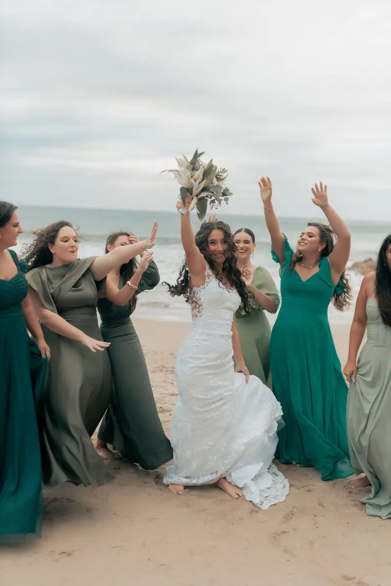 Bride celebrating with bridesmaids on a beach in elegant dresses, capturing a joyous wedding moment.