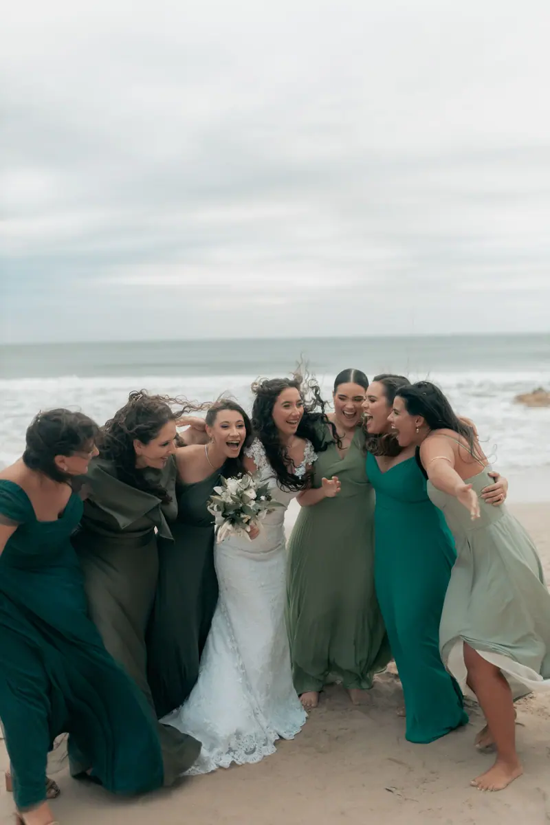 Joyful bridal party in green dresses at a beach wedding, celebrating with laughter.