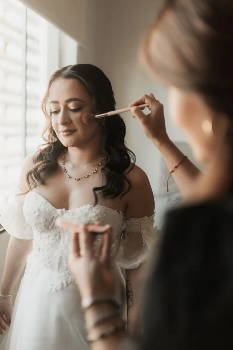 A bride getting her makeup done with a brush in soft, natural lighting by a window.