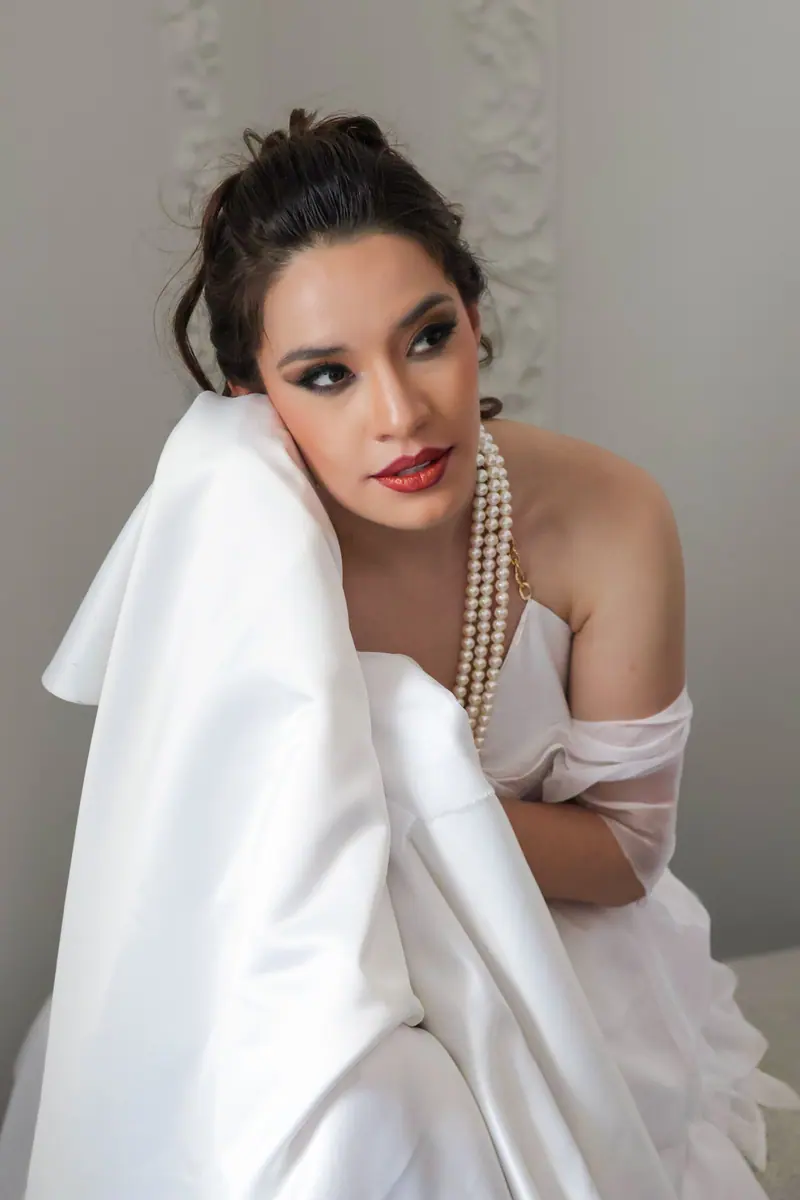 Elegant woman in white gown with pearls posing indoors with sophisticated expression.