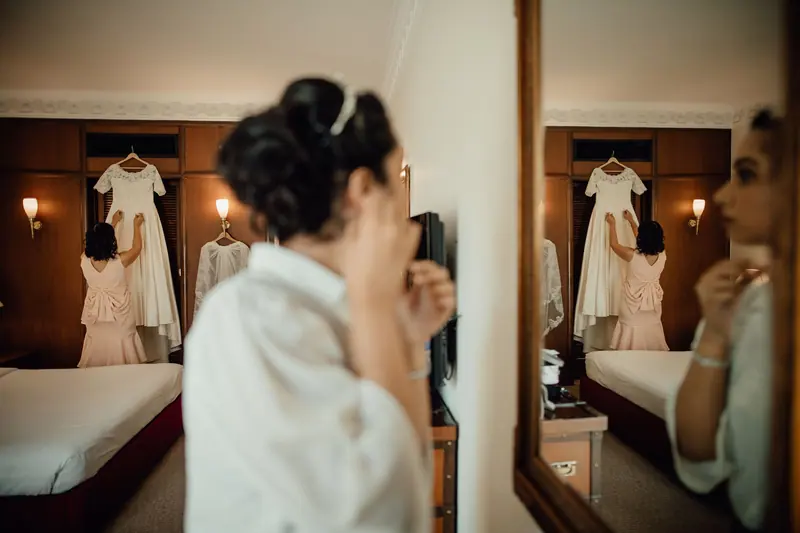 A bride prepares for her wedding day in a hotel room, adjusting her dress in front of a mirror.