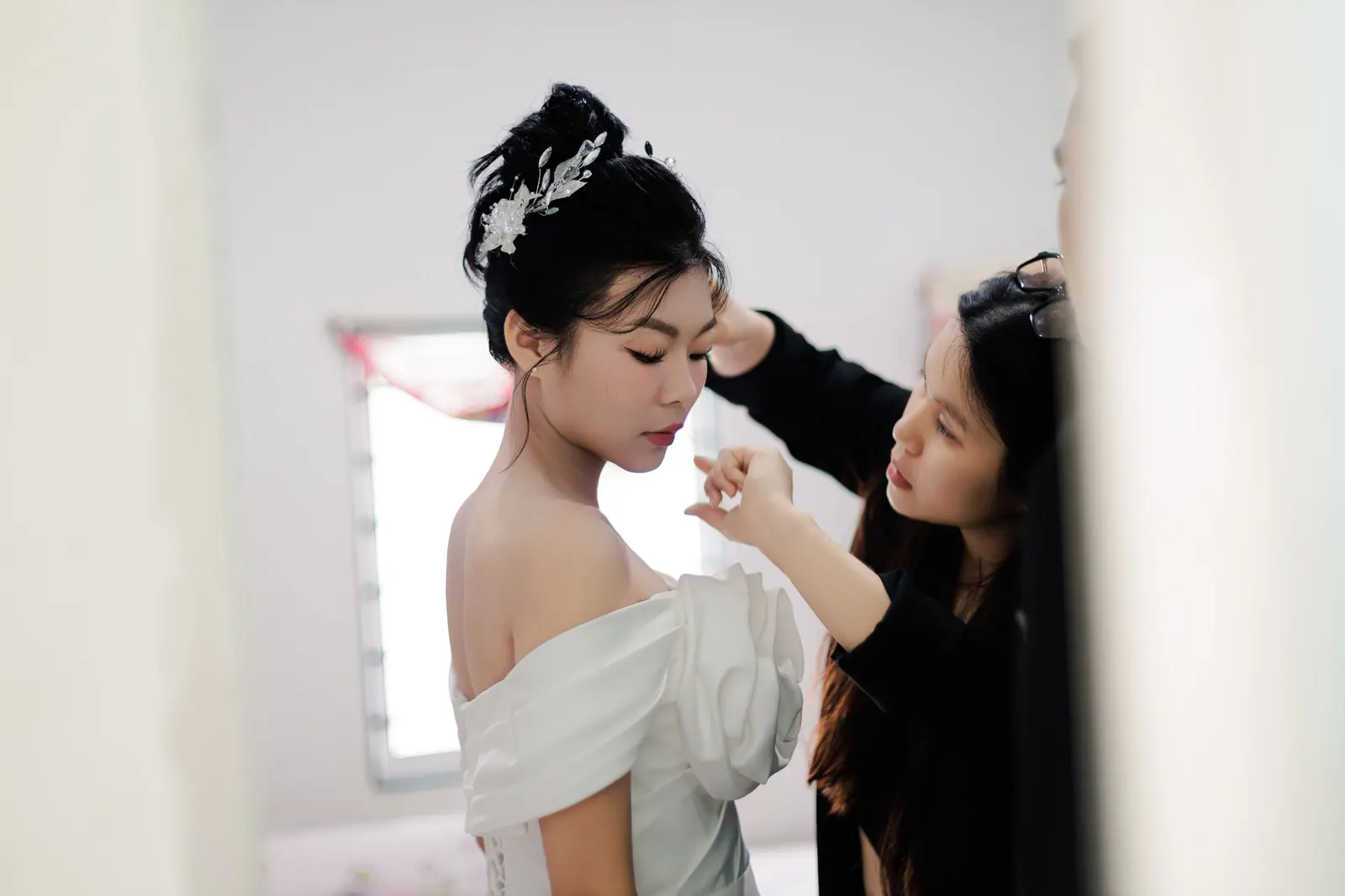 A bride in a white dress being assisted by a makeup artist indoors, showcasing elegance and preparation.