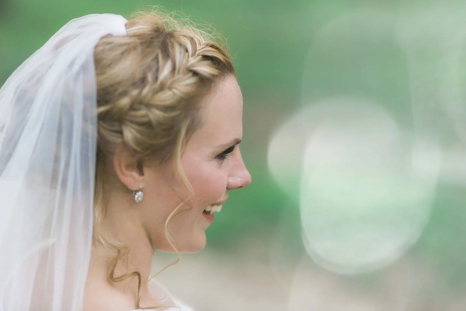 Smiling bride in profile with a braided style and veil against soft green bokeh