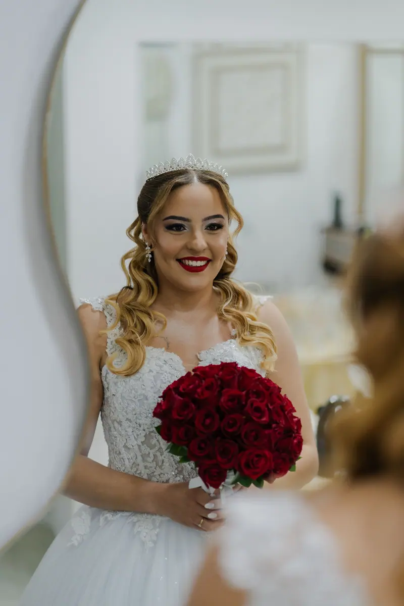 Bride with a bouquet of red roses smiling in a mirror reflection, preparing for her wedding day.