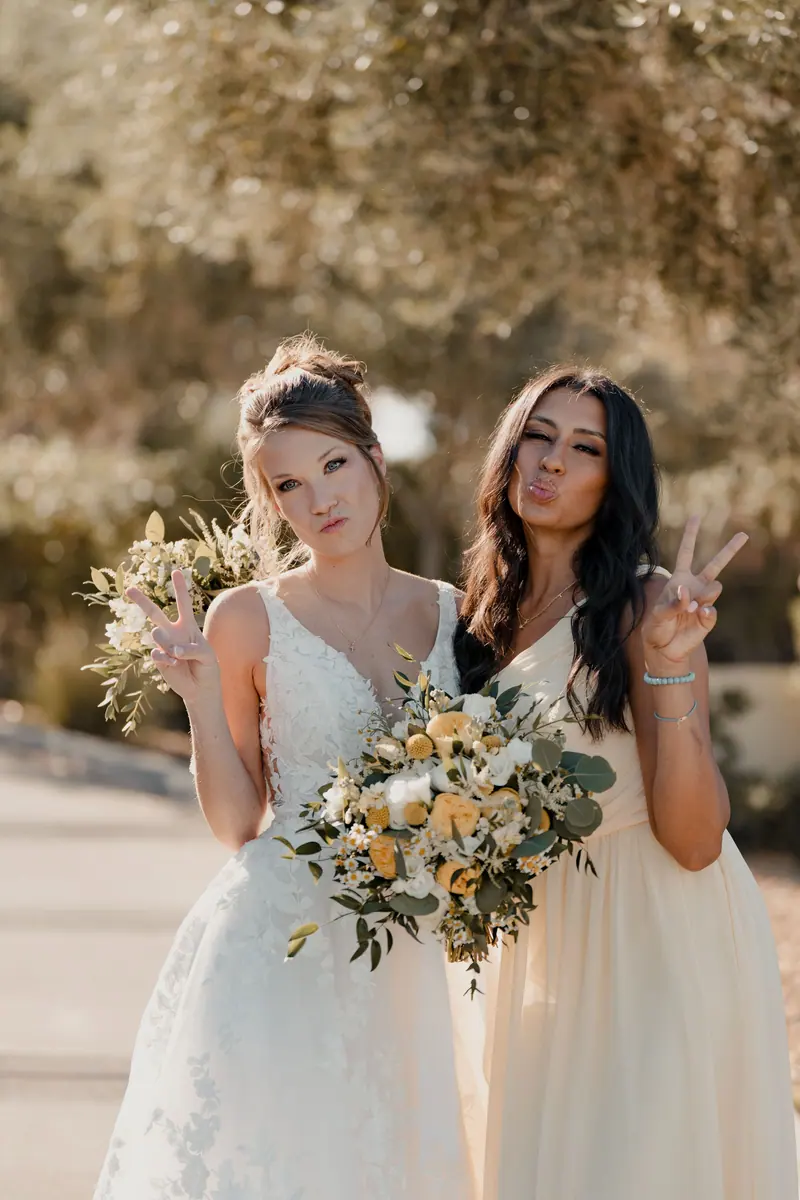 Two women posing playfully at a sunlit outdoor wedding.