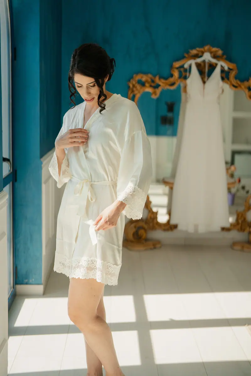 Bride wearing a silk robe stands beside her wedding dress in a sunlit room.