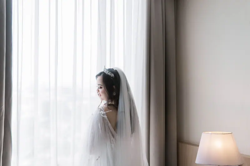 A radiant Asian bride stands gracefully by a sunlit window, exuding elegance.