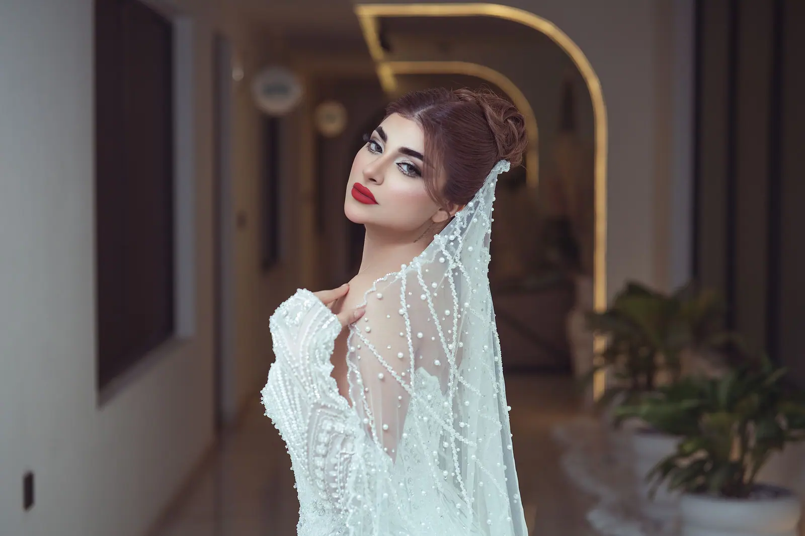 Bride with a sculpted auburn updo, crystal-beaded veil and red lip glancing back down a softly lit hallway