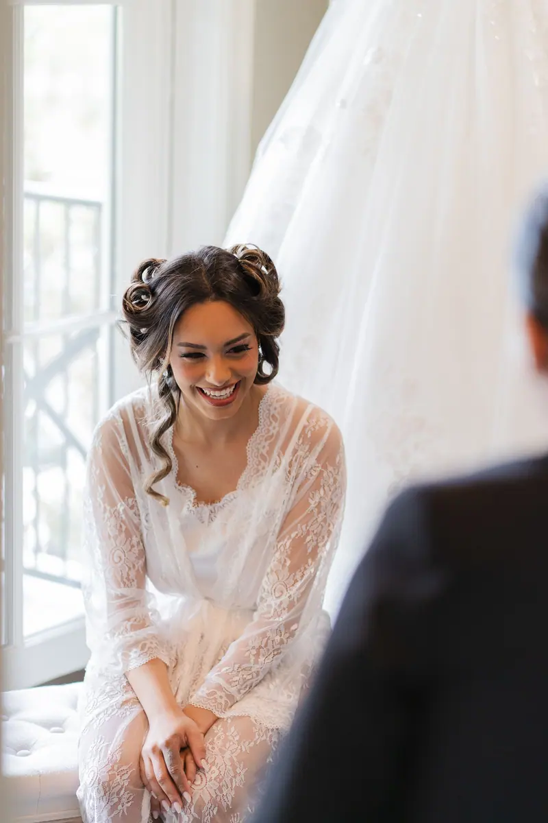 A joyful bride in a white lace robe and hair rollers preparing for her wedding day indoors.