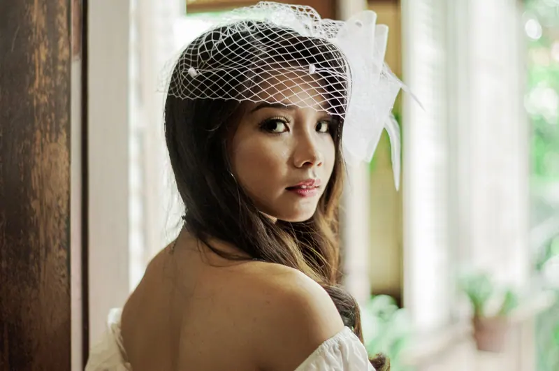 A beautiful bride gazes over her shoulder, dressed elegantly with a white veil, indoors near a window.