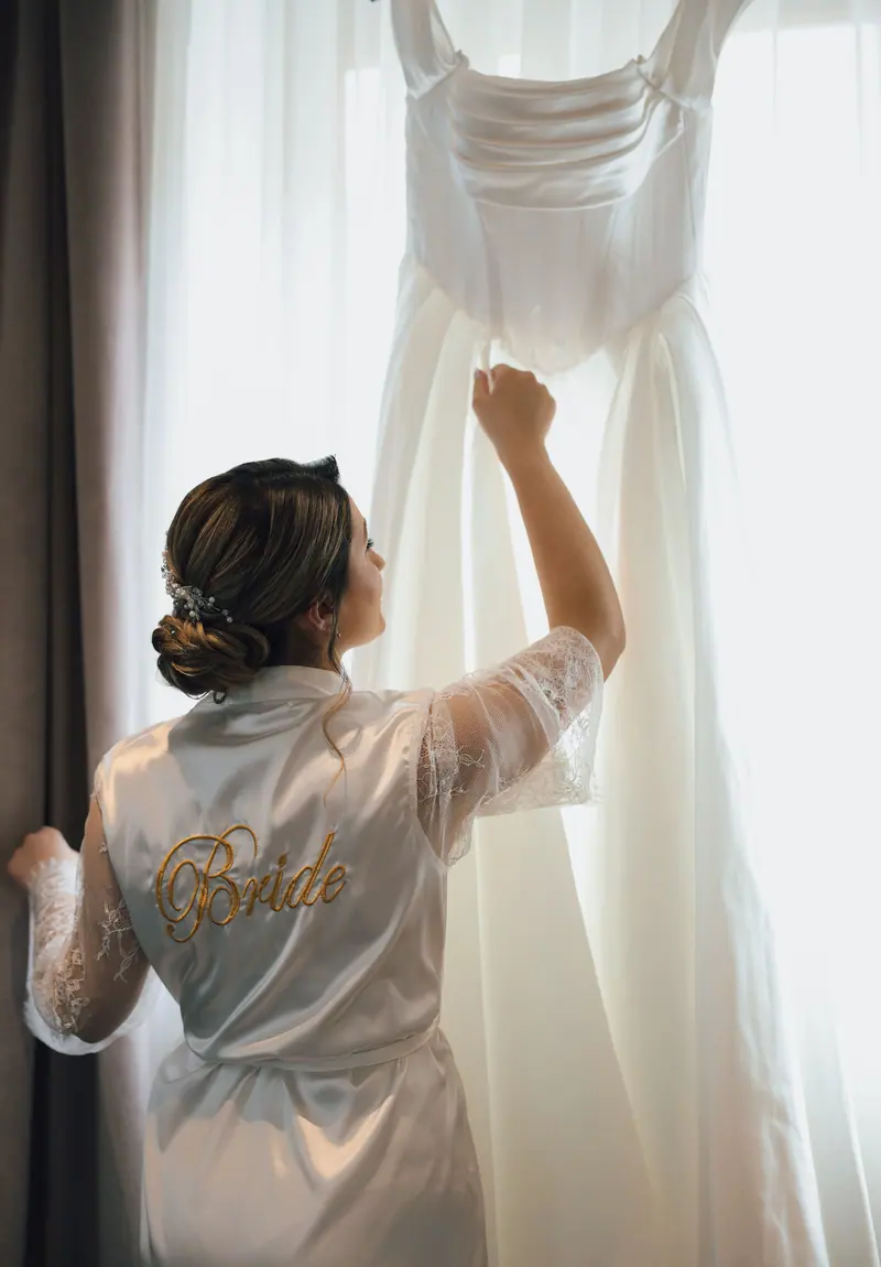Bride in satin robe looking at her wedding dress by a window, exuding elegance and anticipation.