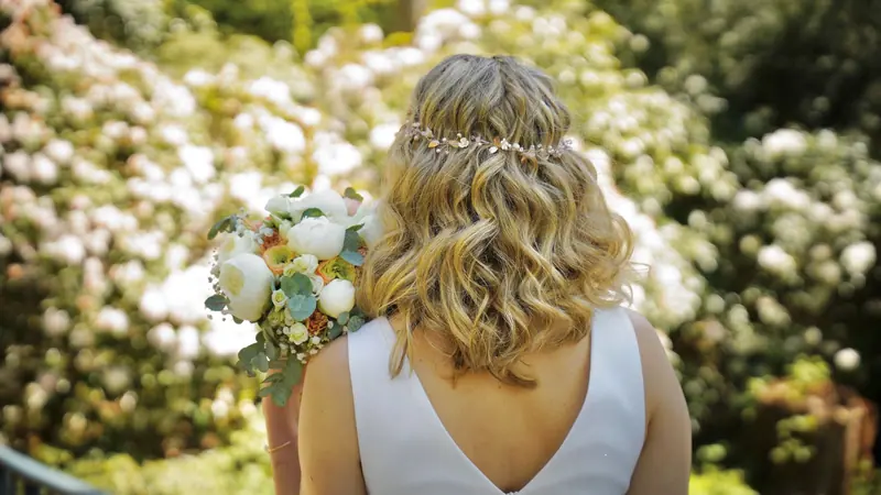 Bride with floral bouquet standing in a blooming garden, showcasing her elegant hairstyle.