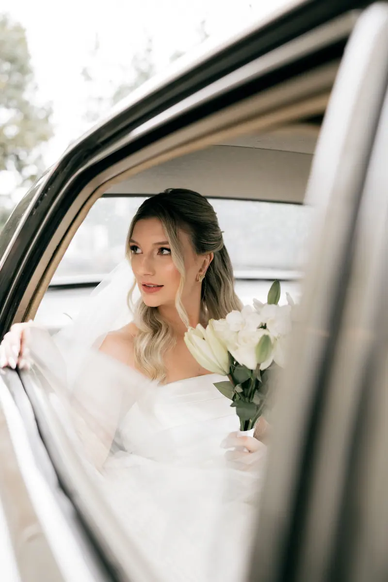 Elegant bride in wedding dress holding a white lily bouquet, looking out from car window.