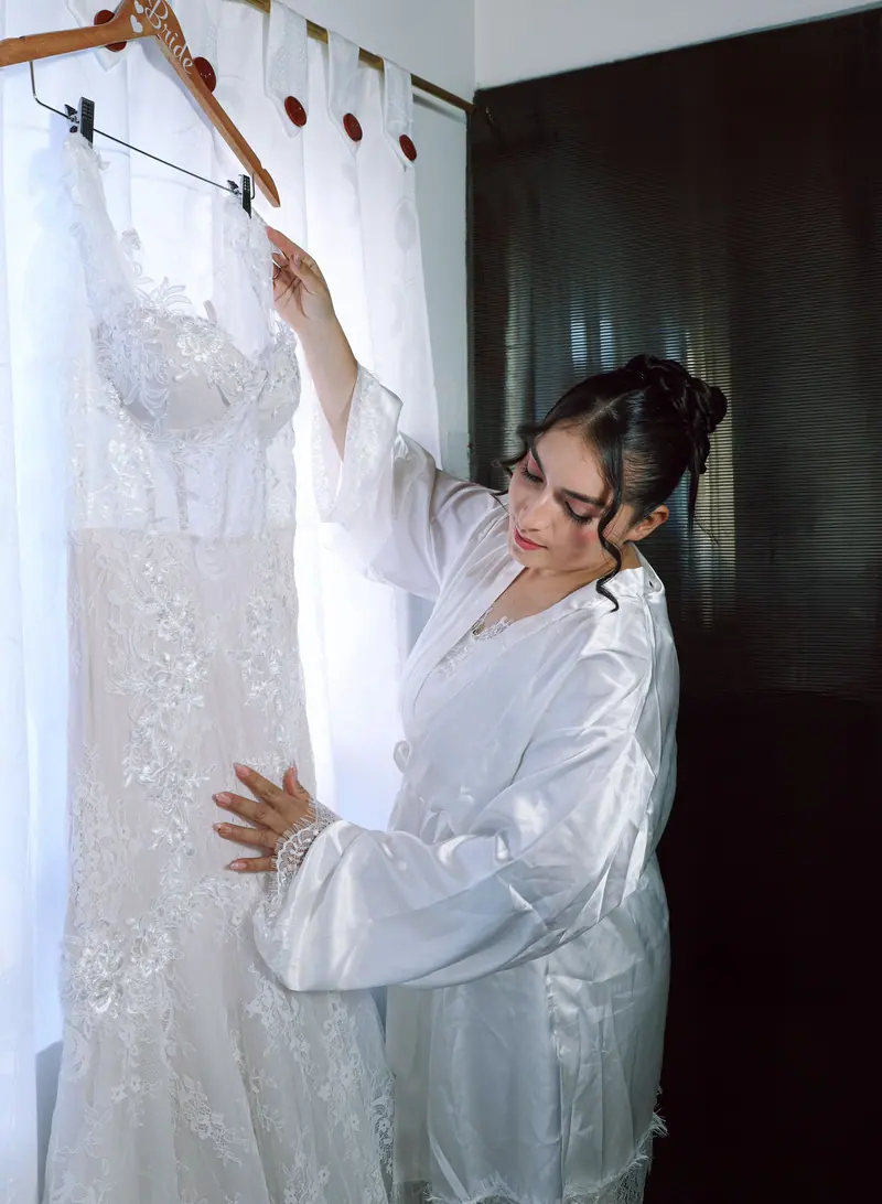 Bride in white robe admires her lace wedding dress by window light.