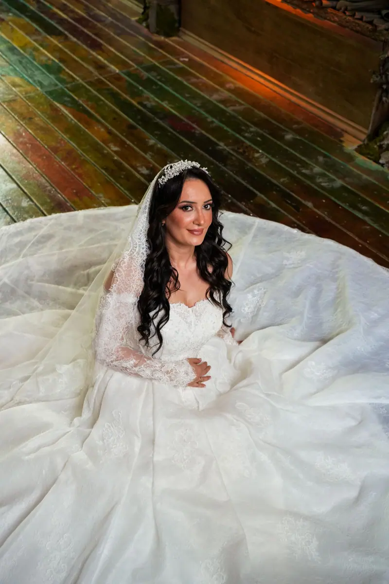 A beautiful bride in a lace gown sits gracefully on a wooden floor, exuding elegance and charm.