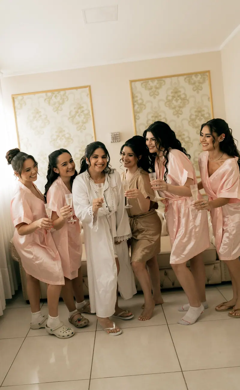 Bridesmaids and bride smiling and celebrating in pastel robes indoors with champagne glasses.