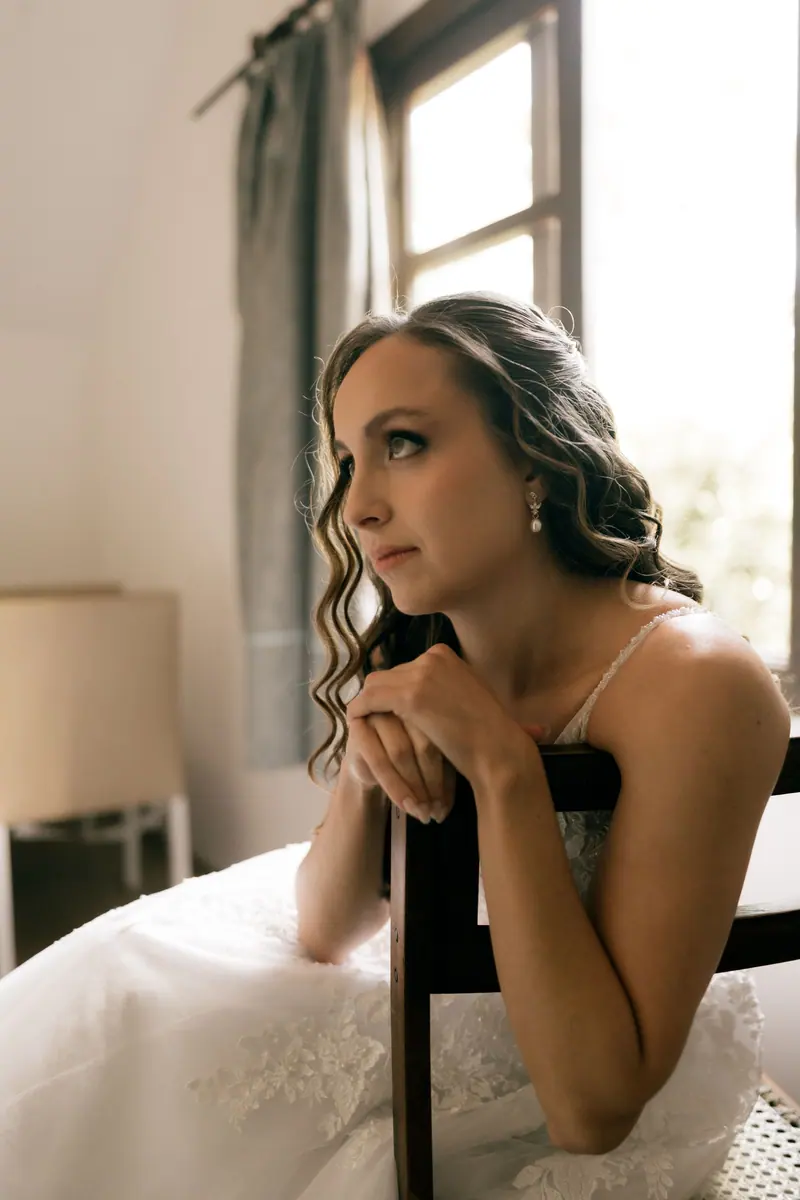 A bride in a white gown gazing thoughtfully out of a window, bathed in soft daylight.