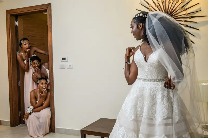 Bride shows off her wedding gown to excited bridesmaids, capturing a joyful and elegant moment indoors.